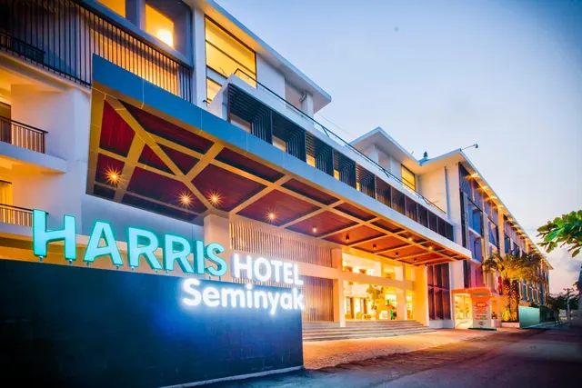 Exterior of the Harris Hotel Seminyak at dusk, with illuminated signage on the front wall above a covered entrance with steps.