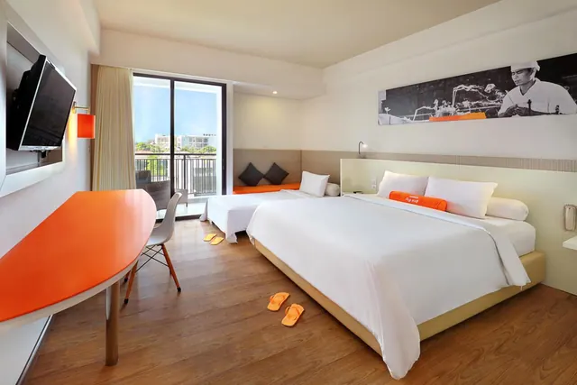 A bedroom with a large double bed, a second bed, an orange desk and chair, a wall-mounted television, and a sliding door onto a balcony overlooking trees and buildings.