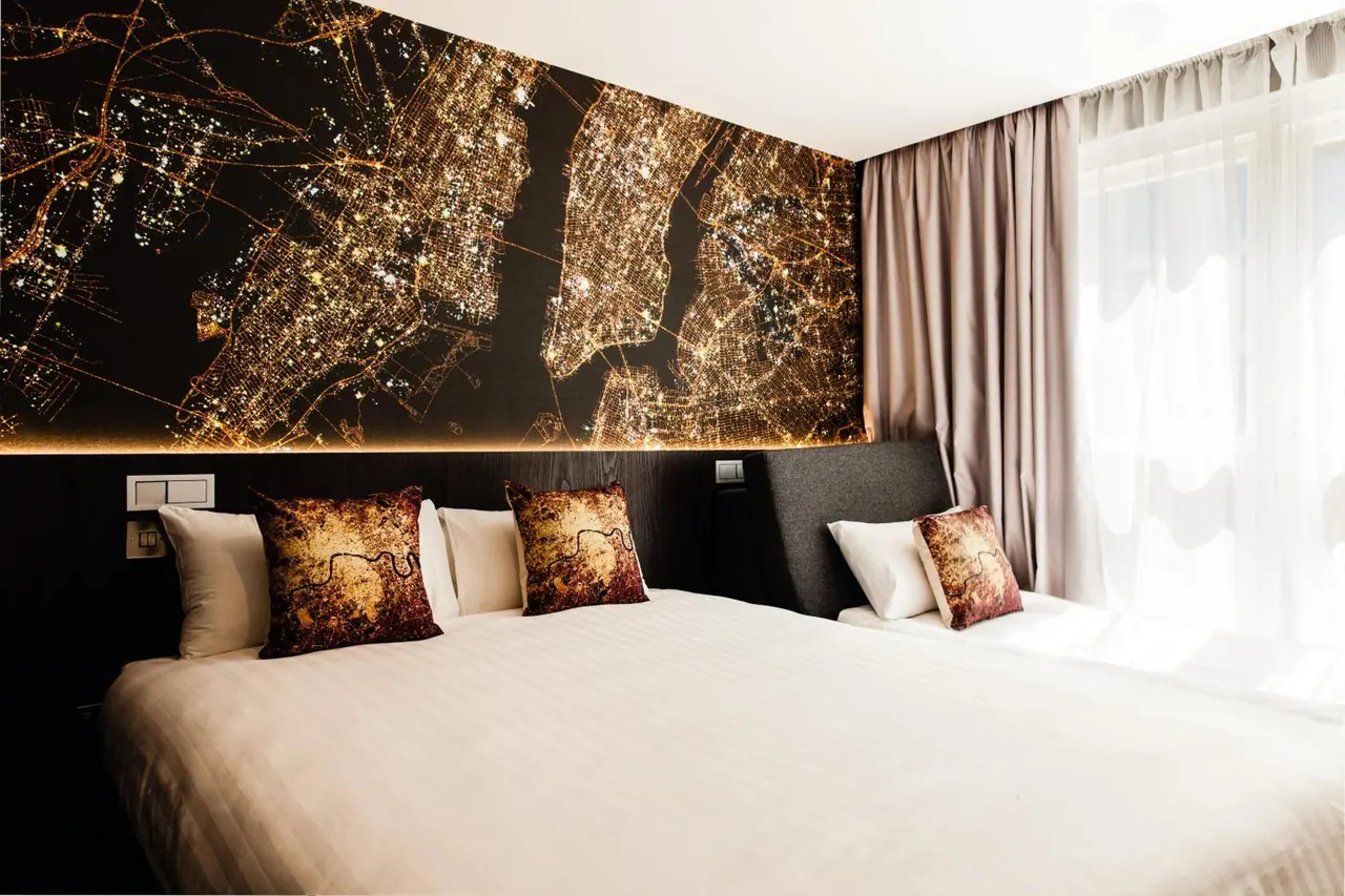 A hotel bedroom with two beds made up in white linen, a night-time city lights map mural on the wall behind them, and sheer curtains over a window on the right.
