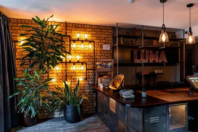 A reception area with an exposed brick wall lit by Edison bulbs, potted plants, a metal-fronted reception desk, and a lit LUMA sign on steel shelving.