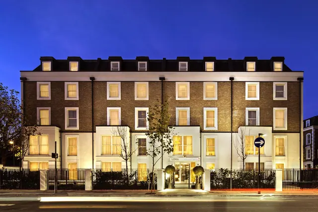 The front of a brick and white stucco hotel building is lit up at dusk under a deep blue evening sky, with topiary plants beside the entrance.
