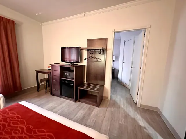 A hotel bedroom with a flat-screen TV on a wooden dresser holding a small fridge and safe, a small desk with a stool, a hanging clothes rail, and a bed with a red spread in the foreground.