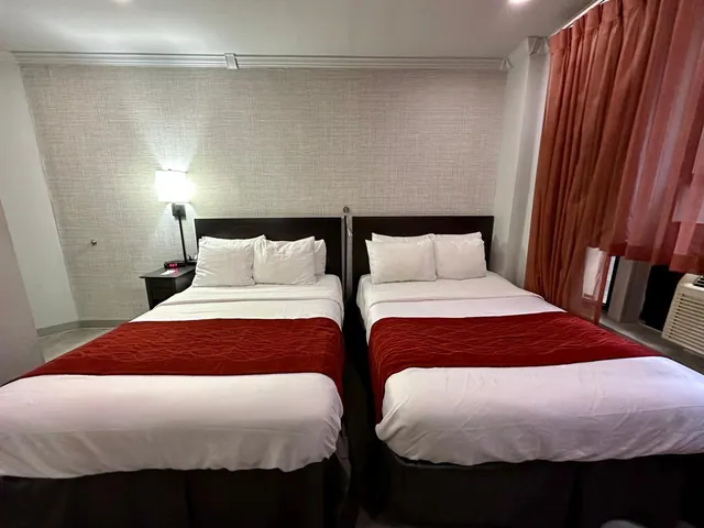 Two made-up double beds with red throws and white pillows sit in a room with orange curtains and a window air conditioner.