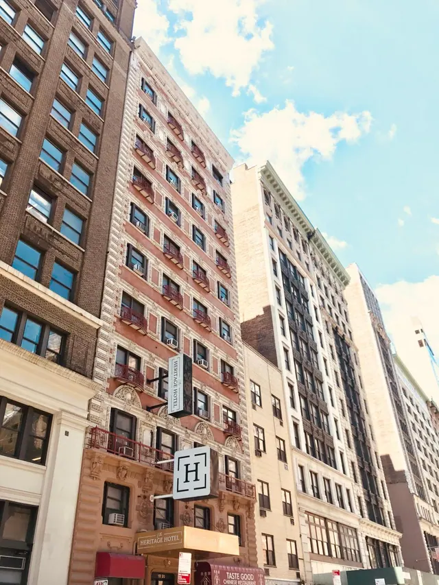 Upward view of the Heritage Hotel facade with its ornate brick and stone front, window air conditioners and neighbouring high-rises under a partly cloudy sky.