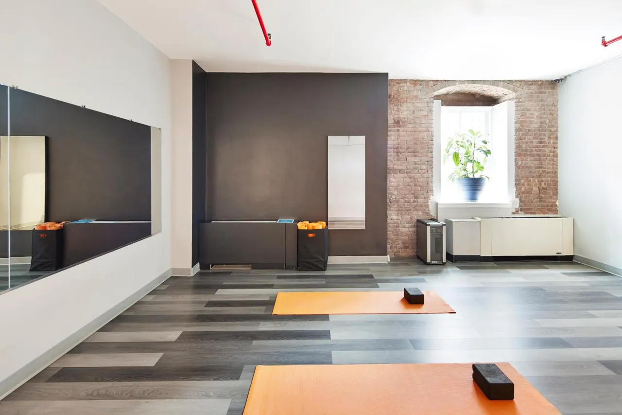An empty studio with orange exercise mats, black foam blocks, wall mirrors, and an exposed brick wall with a window holding a potted plant.