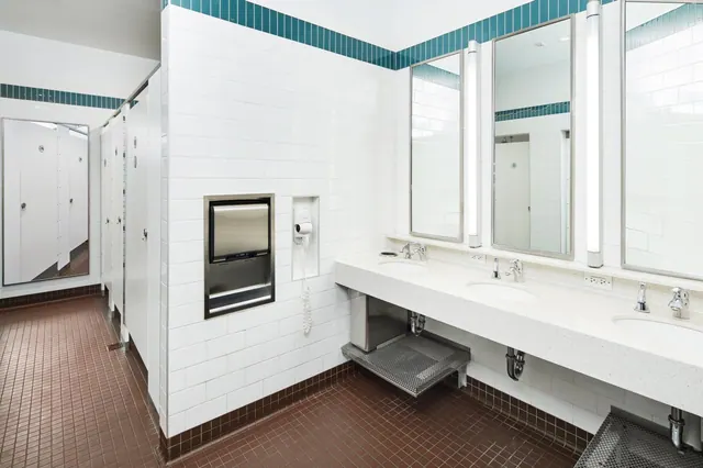 A public restroom with a long white countertop holding several sinks, mirrors above, white tiled walls with teal trim, and stall partitions on the left.