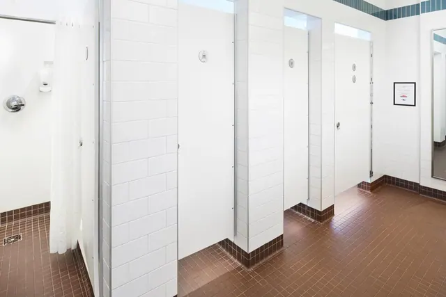 A tiled communal bathroom with white stall partitions, a curtained shower on the left, and a mirror on the right wall.