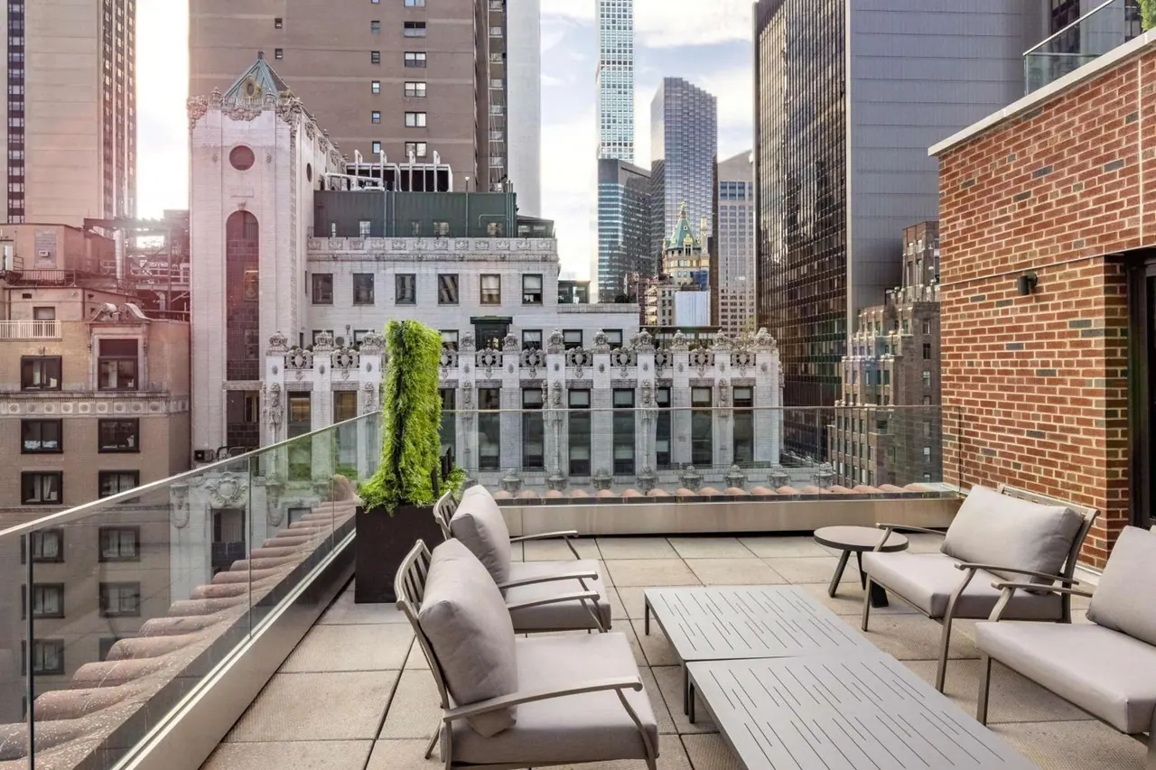 A rooftop terrace with grey lounge chairs, a low table and a glass railing overlooks ornate historic and modern skyscrapers under a partly cloudy sky.