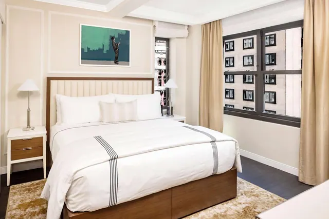 A made king bed with white linens sits between two bedside lamps, beside a window overlooking a neighbouring building.