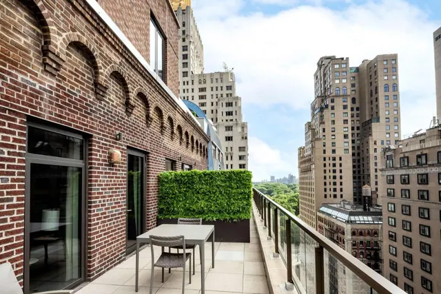A furnished terrace with a table and chairs, a green hedge planter and a glass railing, overlooking high-rise buildings and a band of green trees.
