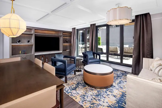 A living area with a dining table, built-in shelving holding a television, two armchairs, a round ottoman and a sofa, with floor-to-ceiling windows onto a terrace.
