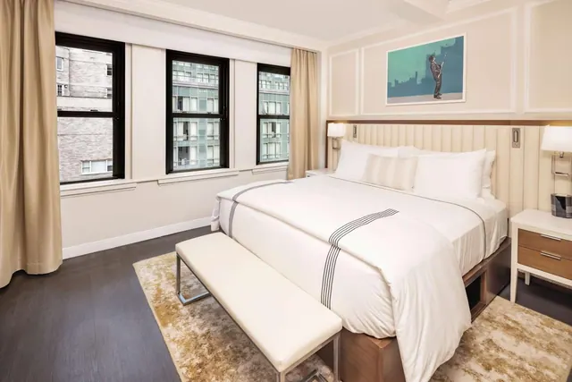 A made king bed with white linen and a cream upholstered bench at its foot, with three windows looking onto neighbouring buildings.