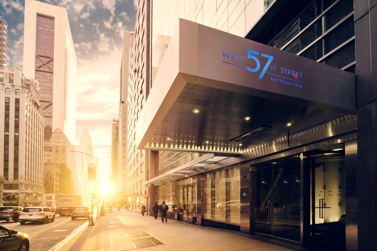 Exterior view of the West 57th Street by Hilton Club entrance under a canopy sign, with two pedestrians and parked cars on the street in low warm sunlight.