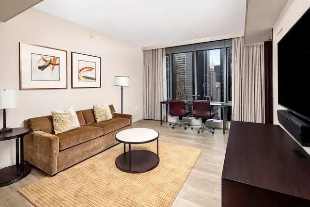 A living room with a brown sofa, a round coffee table, floor lamps, a work desk with two chairs, and a wall-mounted television, with tall windows showing high-rise buildings outside.