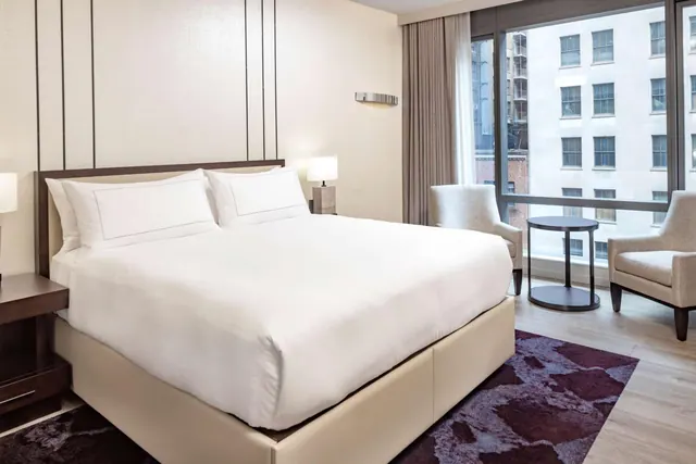 A made king bed with white linens stands beside a large window with two armchairs and a round side table, with a building facade visible outside.