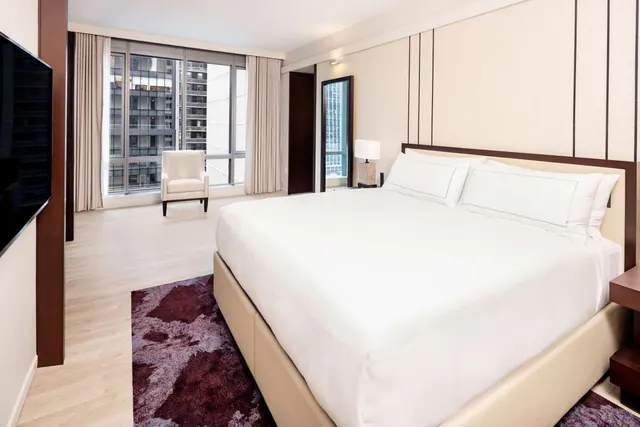 A bedroom with a king bed made with white linens, an armchair by floor-to-ceiling windows overlooking high-rise buildings, and a wall-mounted television.