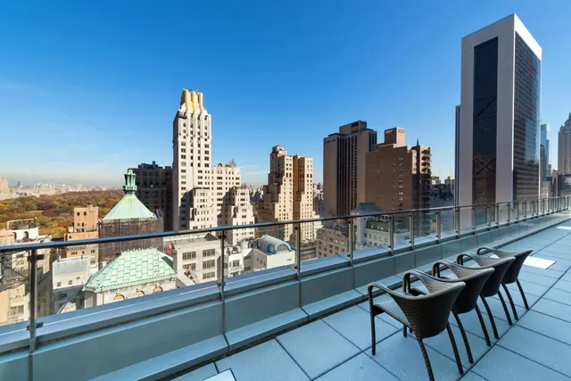 A rooftop terrace with wicker chairs and a glass railing overlooks a dense city skyline and an area of trees under a clear blue sky.