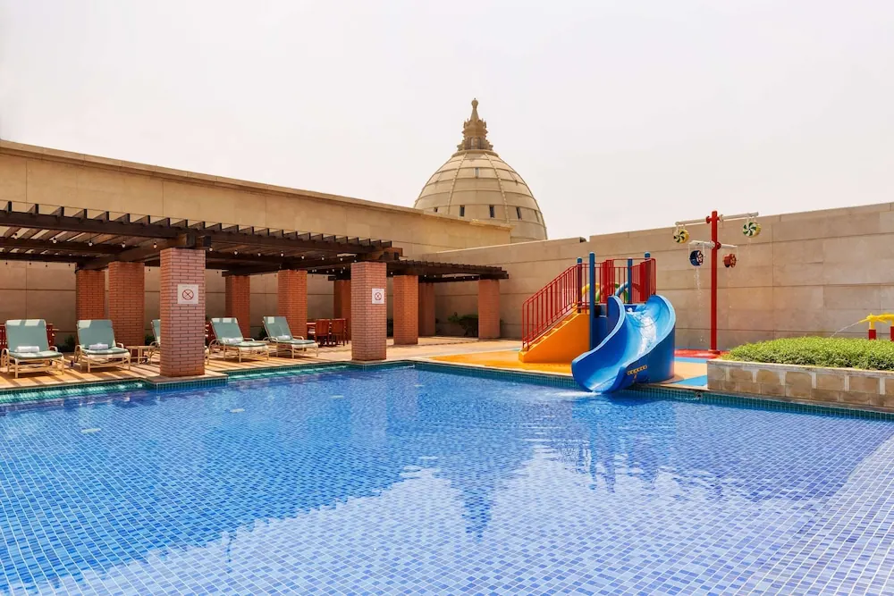 An outdoor pool with blue mosaic tiles, sun loungers under a pergola, and a children's water slide and spray feature beside it.