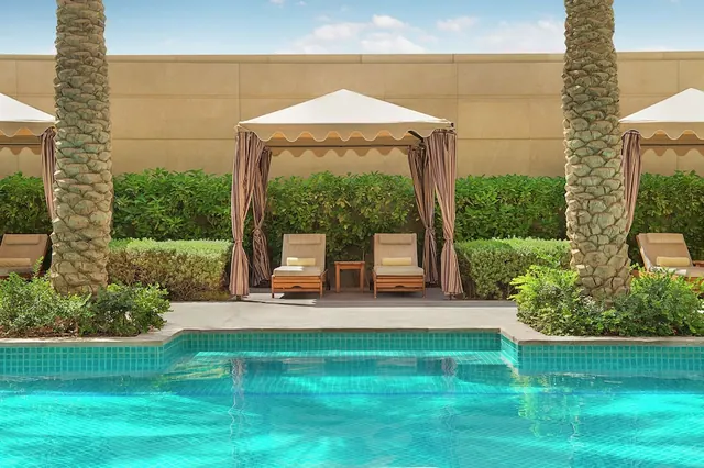 Turquoise pool with tiled edge, two wooden sun loungers under white canopy cabanas with striped curtains, palm trees and green hedges behind a beige wall.