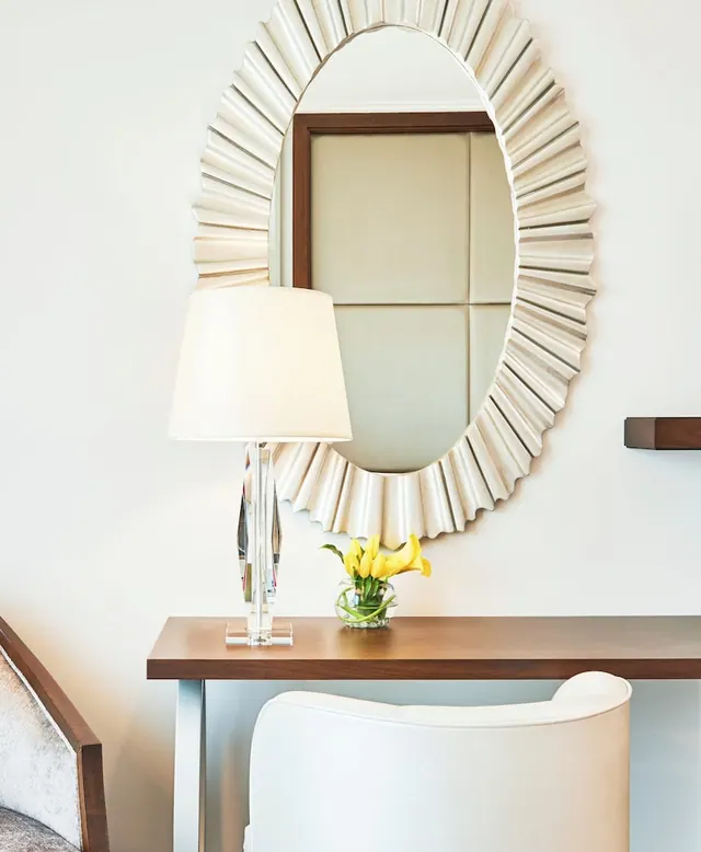 A sunburst mirror hangs above a wooden console table holding a crystal table lamp and a vase of yellow calla lilies, with a white upholstered chair in front.
