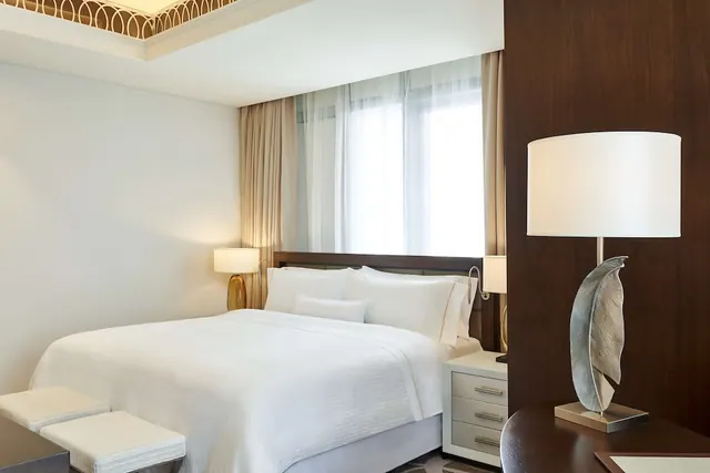 A bedroom with a made king bed with white linen, two bedside lamps, a sheer-curtained window and a dark wood wall panel.