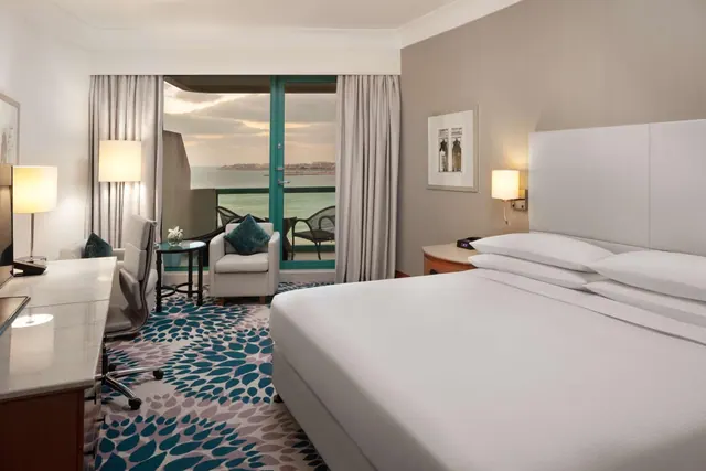 A hotel bedroom with a king bed, a desk, an armchair, and glass doors opening onto a balcony with a view of the sea.