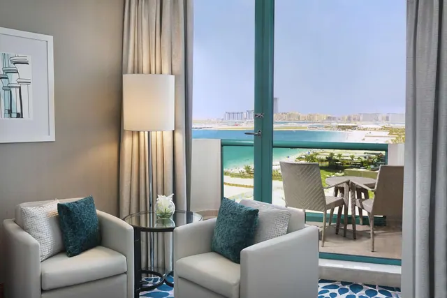 A living area with two white armchairs, a floor lamp and a glass side table, with floor-to-ceiling windows opening onto a balcony with a view of the sea, beach and distant buildings.