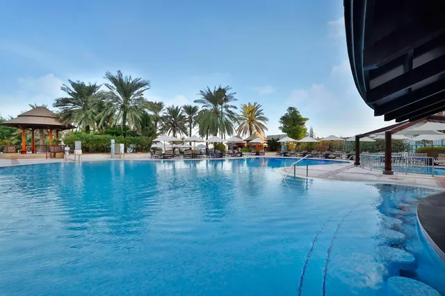 A large outdoor swimming pool with white shade umbrellas and sun loungers beneath palm trees under a clear blue sky.