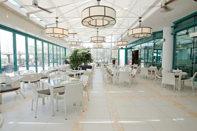 A bright restaurant dining room with white tables and chairs, pendant lights, ceiling fans and large windows looking onto trees outside.