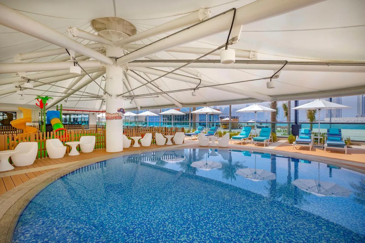 A covered swimming pool with a kids' play area containing a slide, white chairs, blue-cushioned sun loungers and white umbrellas, with city towers visible beyond the glass railing.