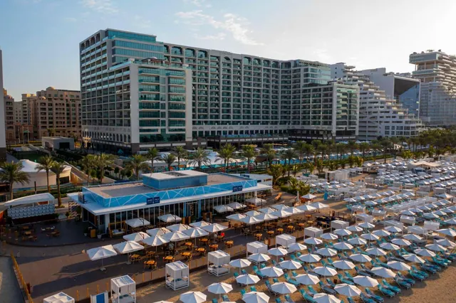 Aerial view of a hotel beach with rows of white umbrellas and sun loungers in front of a tall multi-storey hotel building and a blue-fronted beach restaurant.