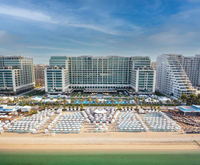 Aerial view of a hotel complex with a sandy beach lined with white umbrellas and loungers, a pool between the buildings, and the sea in the foreground.