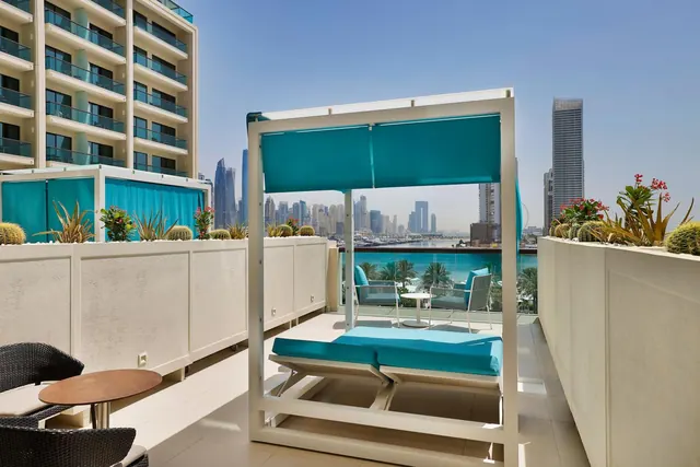 A rooftop terrace with a canopied daybed, wicker chairs, planters with cacti and palms, and a view of high-rise towers across the water.