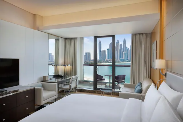 A hotel bedroom with a king bed, a desk, a television and a balcony door looking out over the sea and a row of high-rise towers.