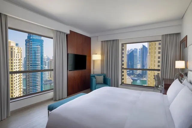 A hotel bedroom with a made king bed, a teal armchair, a wall-mounted television, and two windows overlooking high-rise towers and a waterway.