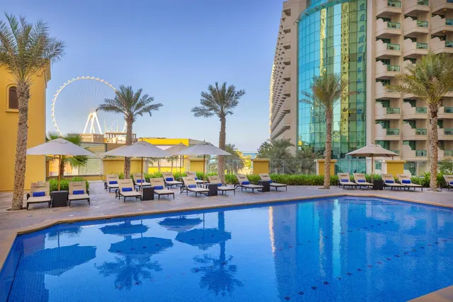 An outdoor pool with blue-cushioned loungers and umbrellas, palm trees, a glass hotel tower, and the Ain Dubai observation wheel in the background.