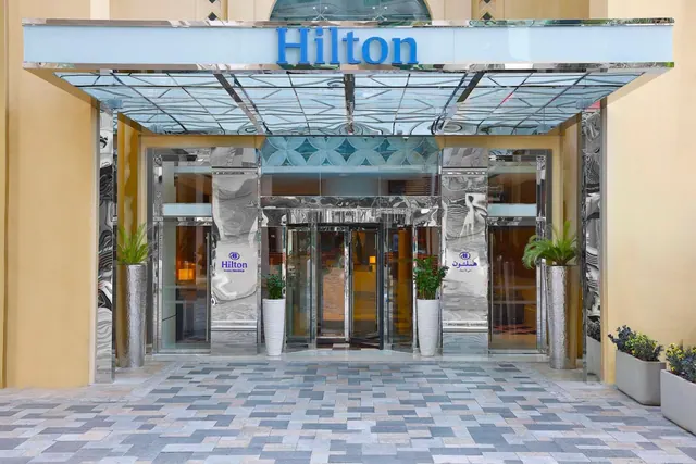 The entrance of a Hilton hotel with a glass canopy, revolving doors, potted plants and a paved forecourt.
