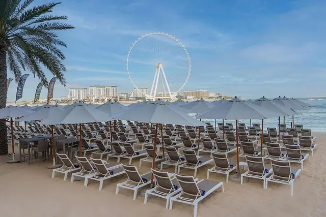 Rows of white sun loungers and grey umbrellas stand on a sandy beach, with the sea and the Ain Dubai ferris wheel in the background.