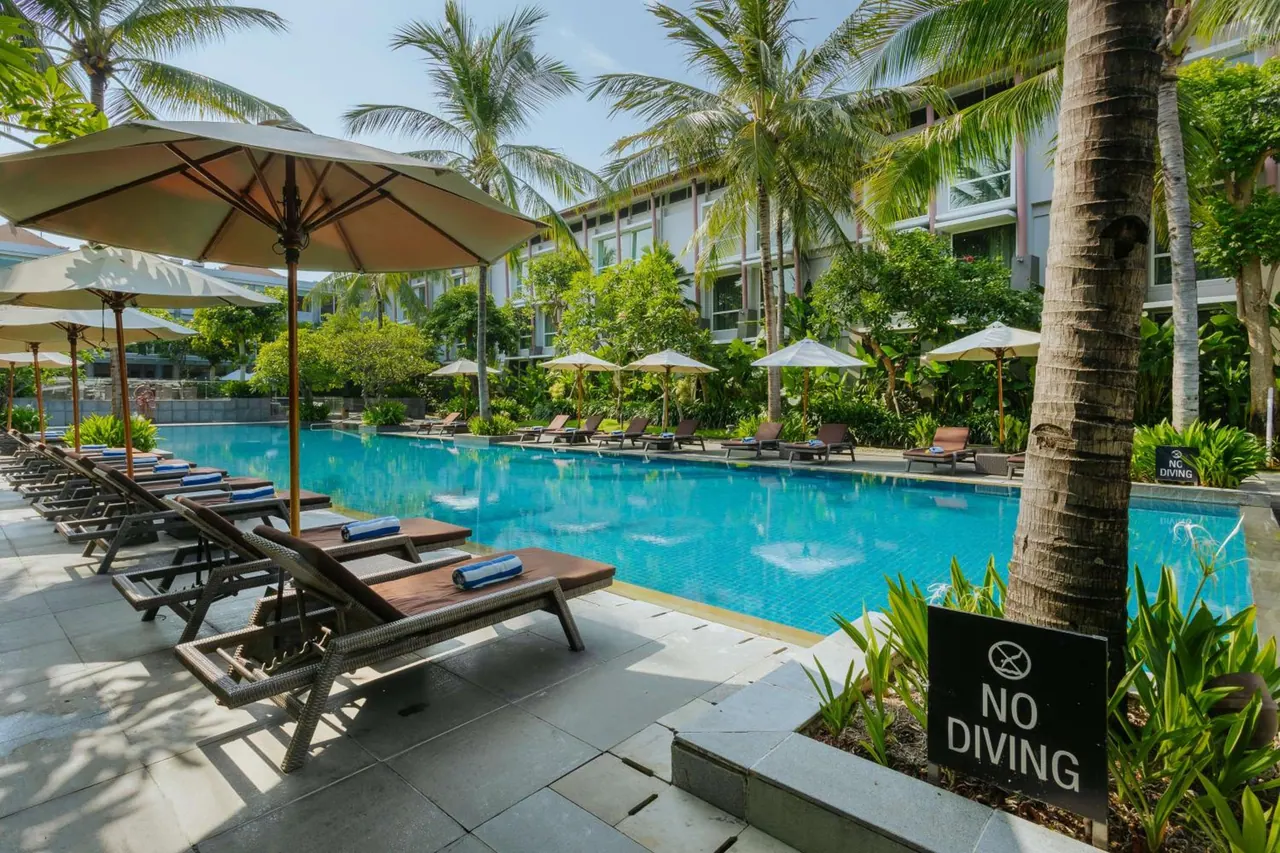 A pool with sun loungers and shaded umbrellas beside palm trees, with a No Diving sign at the pool edge and a hotel building behind.