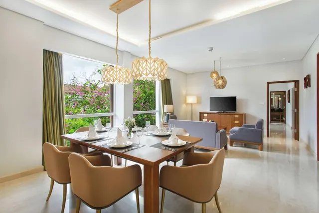 A dining table set for a meal sits beside a grey sofa and a wall-mounted TV in a bright living area, with a window showing trees outside.