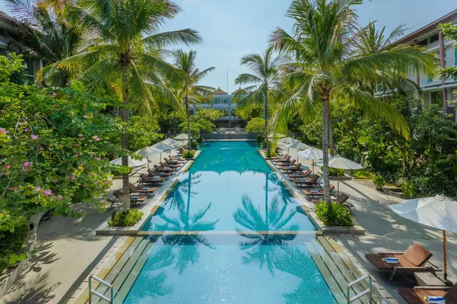 A long lap pool runs between rows of sun loungers and white parasols, with palm trees and tropical planting on both sides.