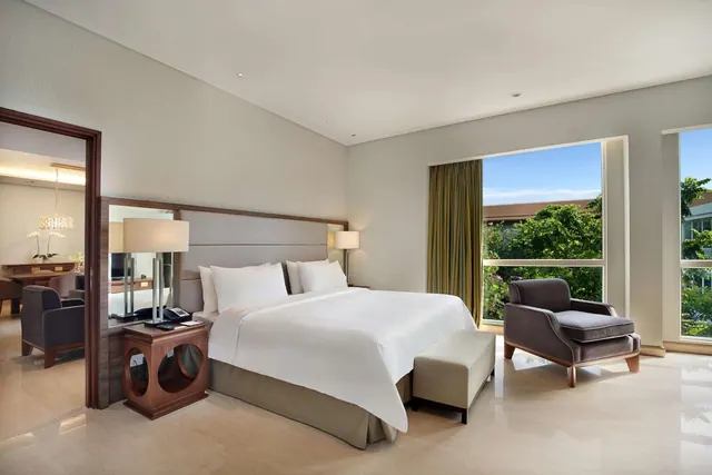 A bedroom with a king bed, a grey armchair and a padded bench at the foot of the bed, with large windows looking out onto green trees.
