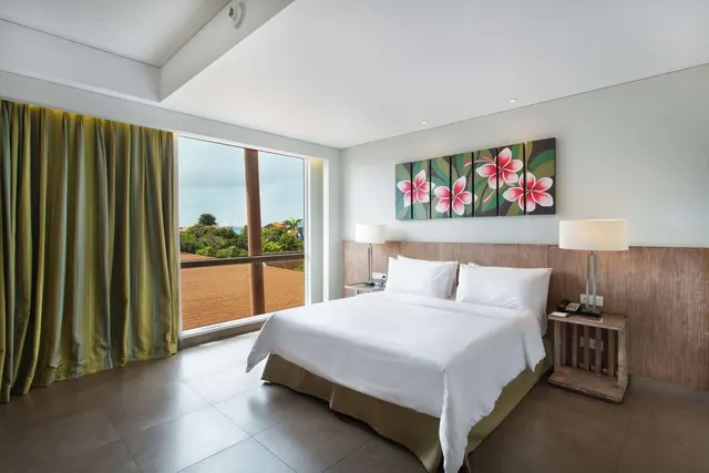 A king bed with white linen sits beside a floor-to-ceiling glass door that opens onto a balcony with a view of trees, and a floral painting hangs above the headboard.