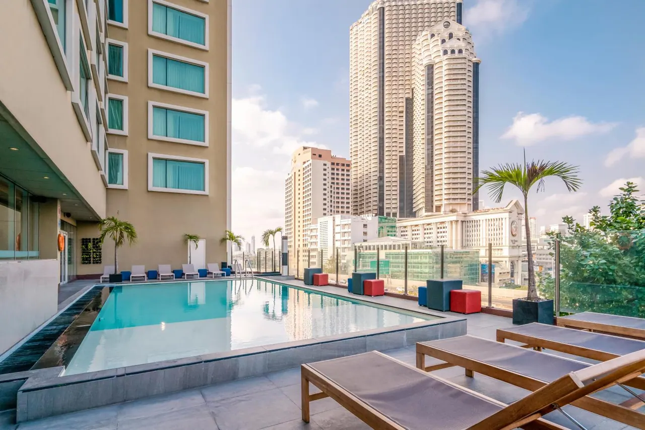 Rooftop swimming pool with sun loungers beside a terrace, with high-rise buildings visible in the city beyond.