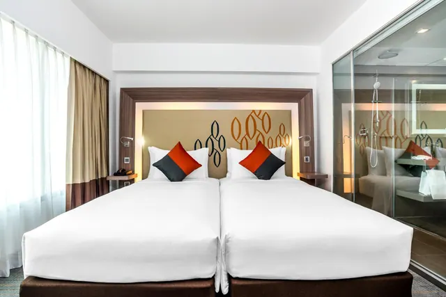 Two single beds with white linen and orange and grey cushions sit in a bedroom beside a glass-walled shower area.
