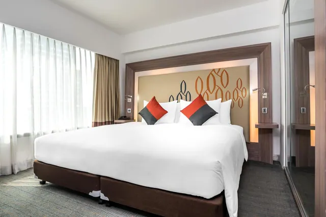 A made bed with white linen and two red and grey cushions sits against a patterned headboard, with sheer curtains at the window and a mirrored wardrobe on the right.