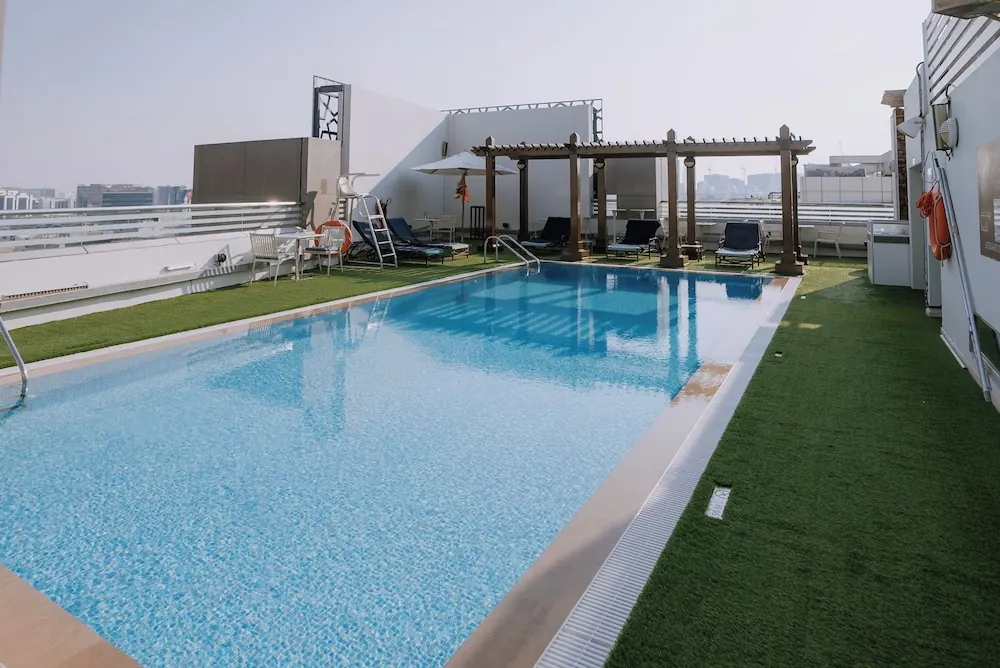 A rooftop swimming pool with artificial grass surrounds, sun loungers, a wooden pergola and a white parasol.