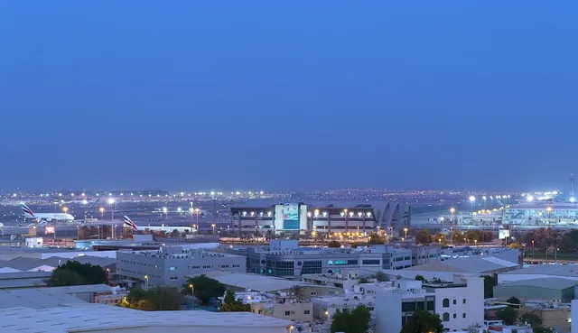 Elevated twilight view over Dubai International Airport with illuminated runways, parked aircraft, a terminal building and low rooftops in the foreground.