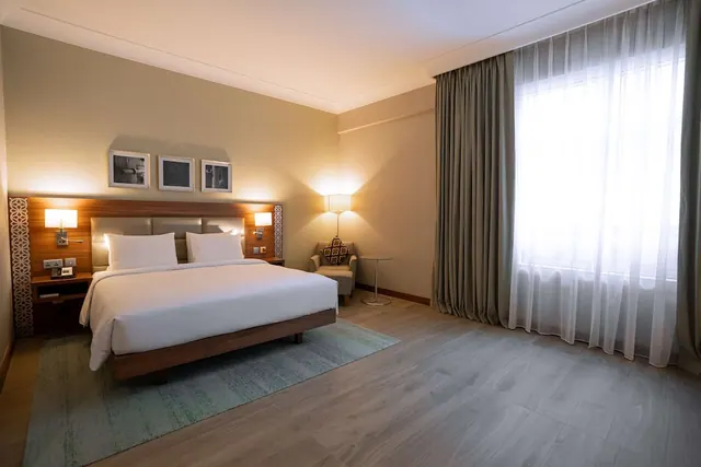 A bedroom with a king bed, wooden headboard, bedside lamps, and an armchair beside a window with sheer curtains.