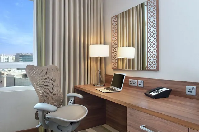 A wooden work desk with a laptop, a telephone, two table lamps and a mirror, with a mesh desk chair beside a window showing city buildings.